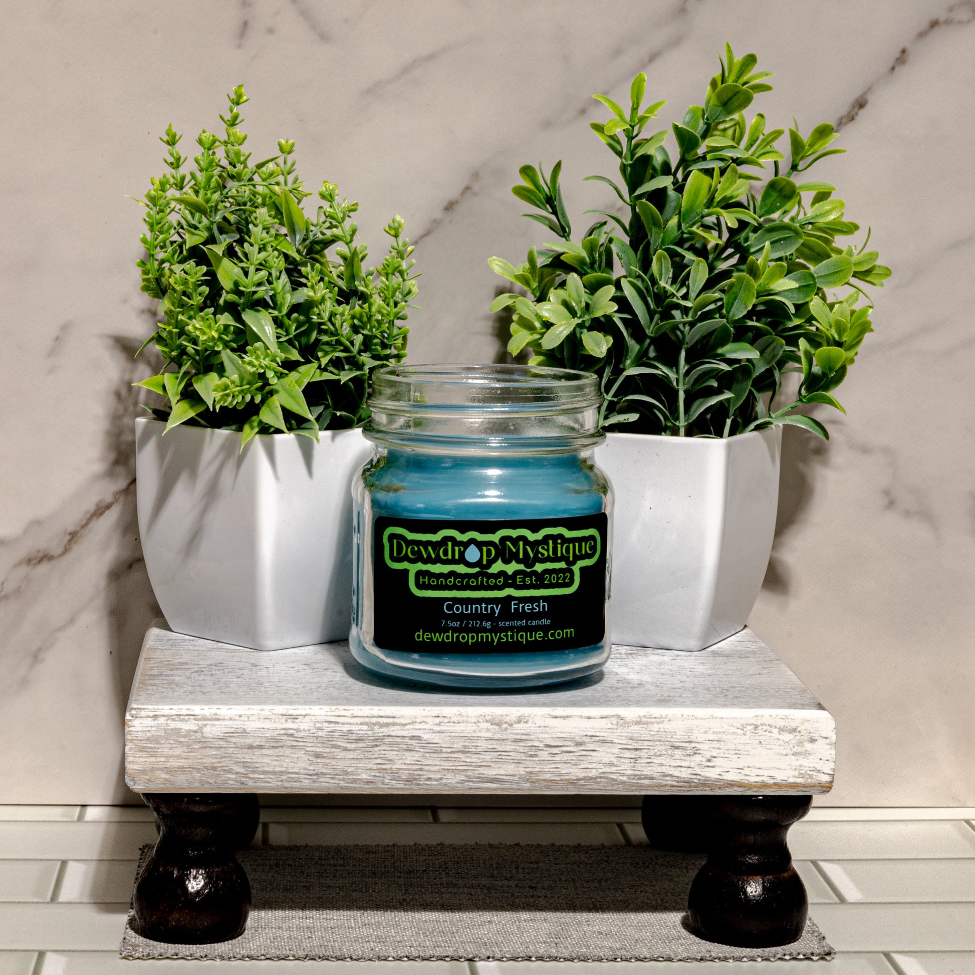 A blue glass candle with a black label that reads 'DEW DROP MYSTIQUE'S CANDLES', placed on a wooden shelf with green plant decor in the background.