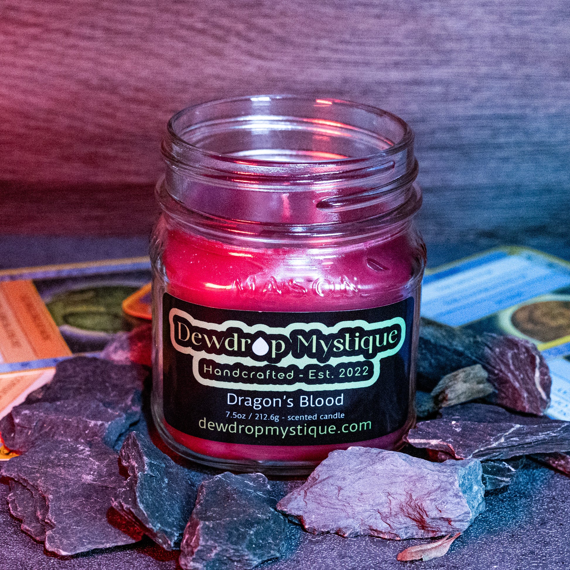 A red candle with a label that reads 'DRAGON'S BLOOD Handcrafted Est. 2022' placed on a dark stone surface, with a blurred background suggesting a mystical or magical theme.