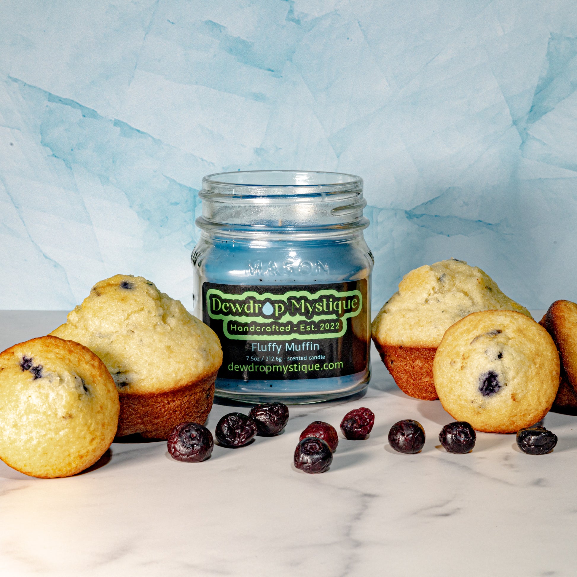 A clear glass jar candle with a label reading 'DEWDROP MYSTIQUE' and 'Fluffy Muffin' surrounded by muffins with blueberries on top.