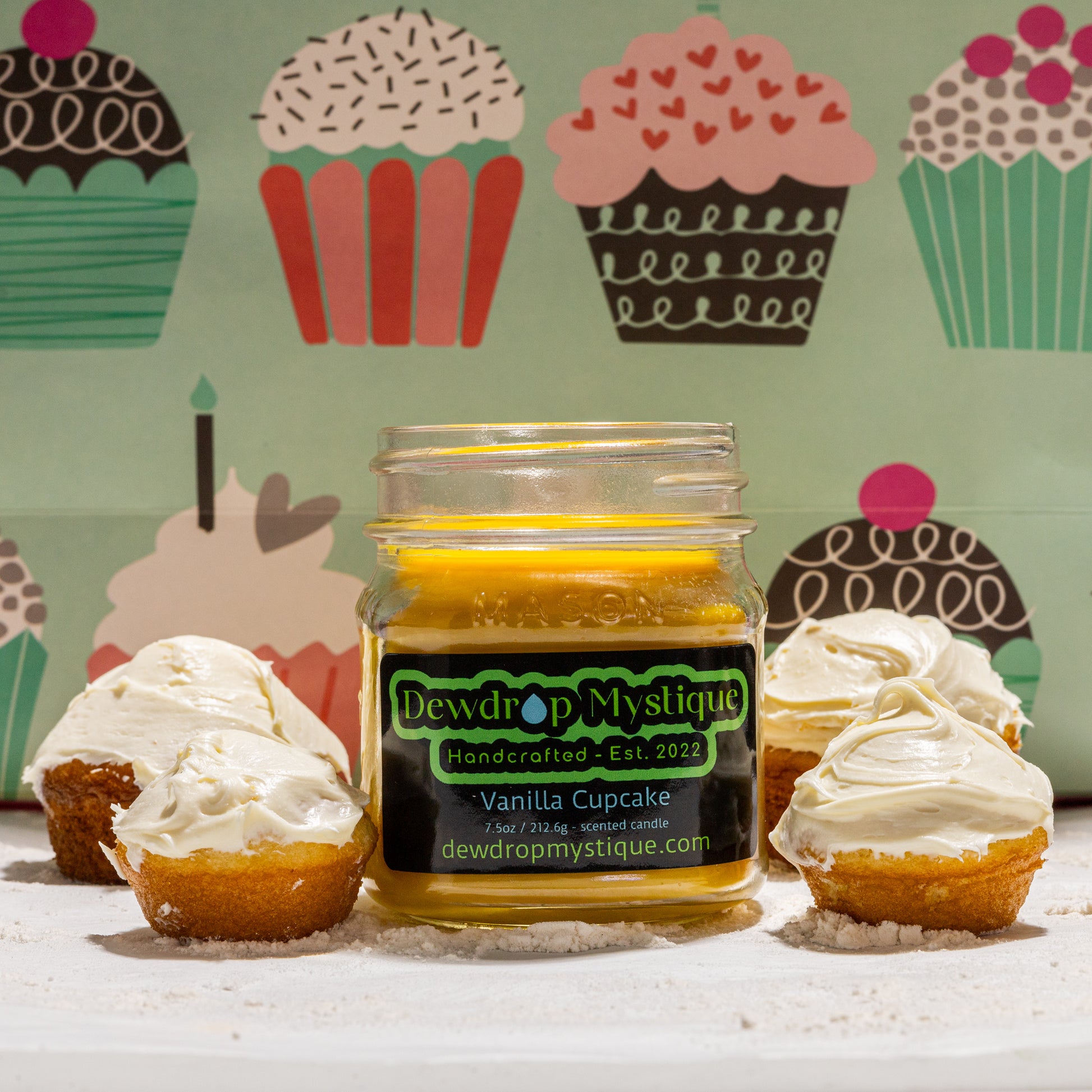 A clear glass jar with a label reading 'Vanilla Cupcake' and a decorative background featuring cupcakes and the Dewdrop Mystique brand logo.