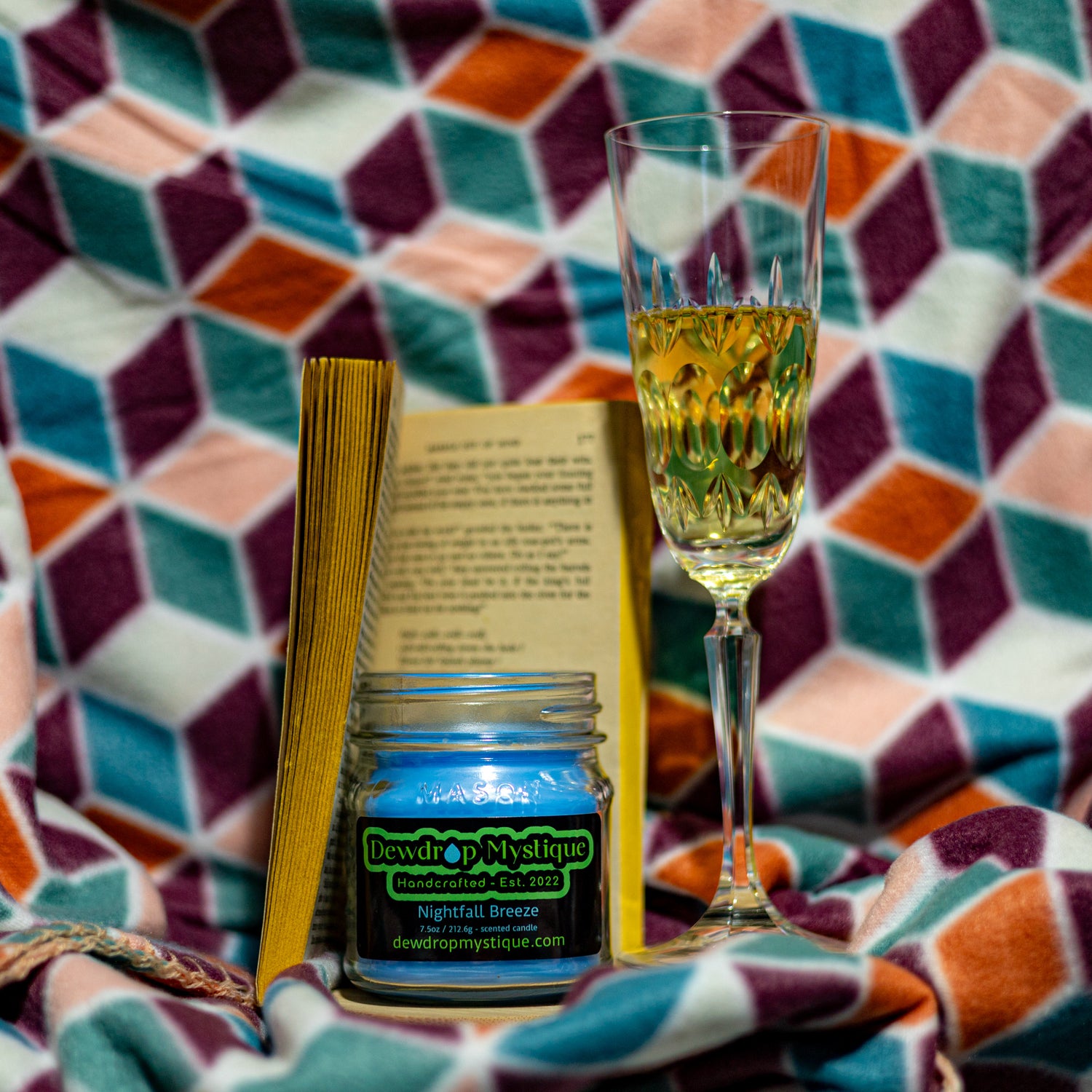 A 7.5oz Nightfall Breeze candle with a blue label, placed on a book, in front of a geometric patterned blanket.